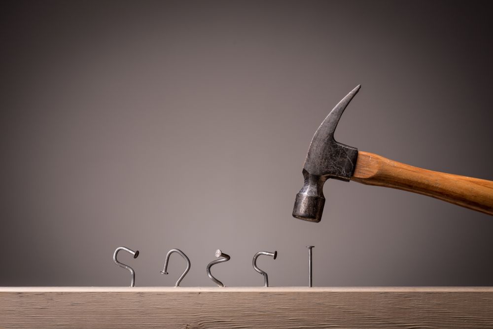 Four Bent Nails In A Plank Of Wood And Hammer Poised To Hammer A Fifth