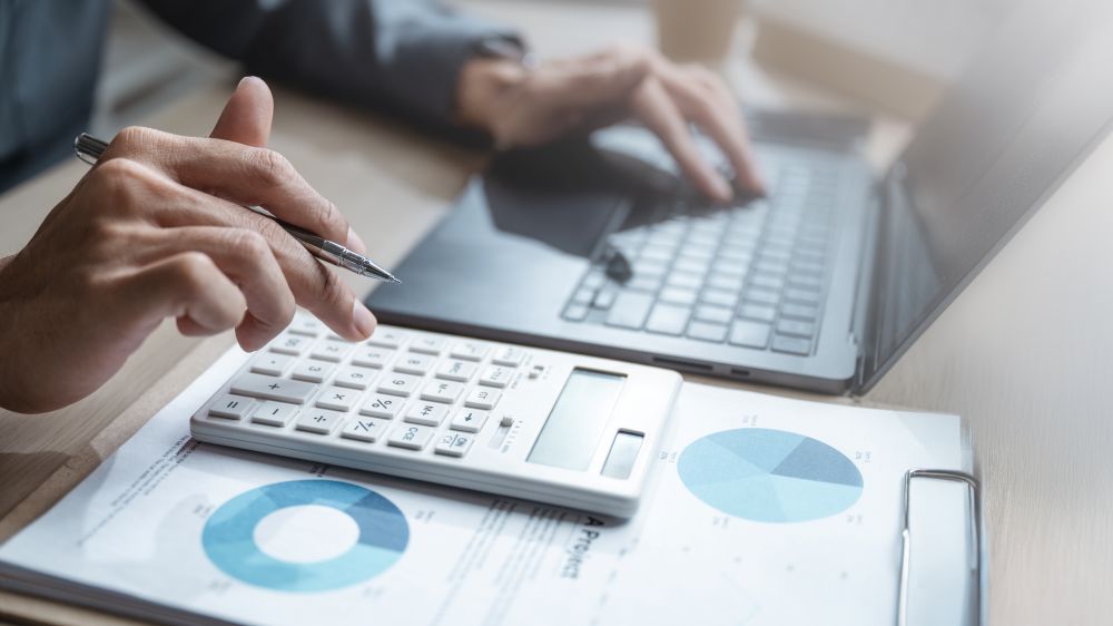 Person with pen typing on laptop and calculator with clipboard and charts
